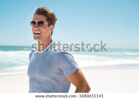 Feliz joven con ropa informal usando gafas de sol y sonriendo en la playa. Un tipo guapo riendo de la playa y mirando lejos. Retrato de un tipo alegre que disfruta de vacaciones de verano con espacio para copiar.