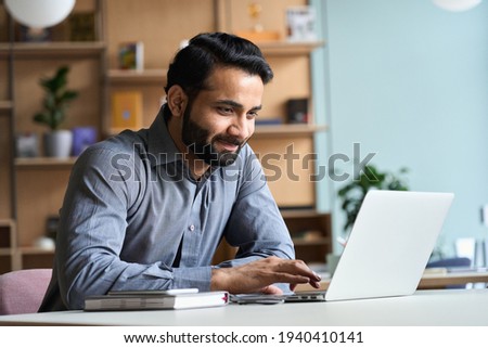 Smiling indian business man working on laptop at home office. Young indian student or remote teacher using computer remote studying, virtual training, watching online education webinar at home office. Stock foto © 