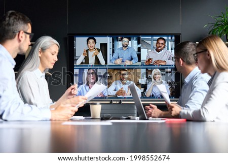 Videoconferencia en línea de corporaciones globales en sala de reuniones con diversas personas sentadas en oficinas modernas y colegas multiculturales en monitor de pantalla grande. Concepto de tecnologías empresariales.