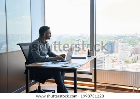 Indian latin successful thoughtful businessman leader thinking of future corporate successful plan ideas sitting at office desk looking at panoramic view window of building of modern corporation.