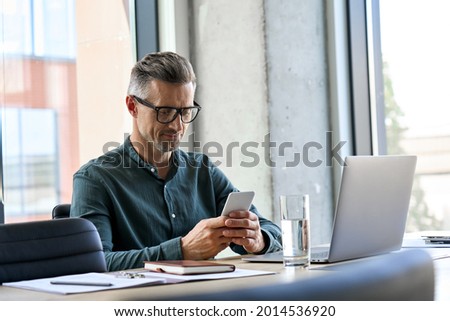 Smiling mature businessman holding smartphone sitting in office. Middle aged manager ceo using cell phone mobile apps and laptop. Digital technology applications and solutions for business development Foto stock © 