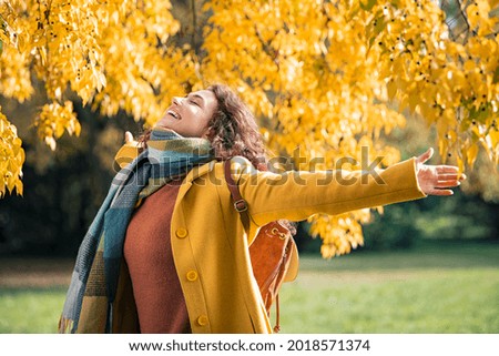Beautiful young woman relaxing at park during autumn season with closed eyes. Happy free natural girl breathing deeply in park with foliage in background. Pretty woman expressing freedom outdoor.