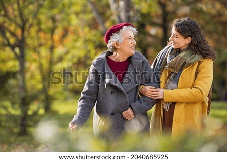 Mujer joven en el parque con ropa de invierno caminando con abuela mayor. Hermosa cuidadora y anciana sonriente caminando en el parque durante el otoño y mirándose entre ellos con espacio para copiar. 