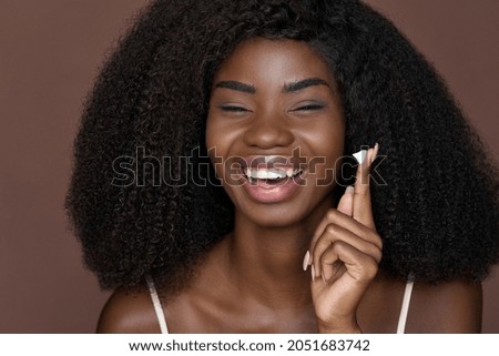 Happy young black woman laughing holding creme on finger applying facial cream isolated on brown background. Cheerful African model advertising moisturizer face skin care product, skincare beauty.