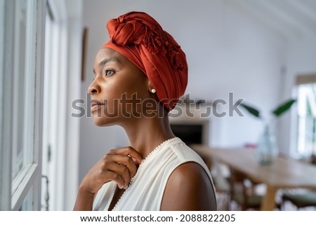 Pensive woman wearing headscarf looking outside window in contemplation. Mature black woman wearing a traditional turban and thinking near window at home. Worried african mature woman with cancer. Foto stock © 