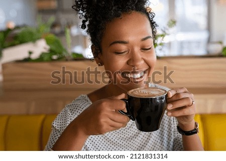 Smiling black young woman smelling freshly brewed coffee with eyes closed in cafeteria. Beautiful african girl smiling while relaxing in a coffee shop. Close up face of girl drinking latte coffee.