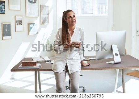 Smiling young female architect leaning on her desk in an office drinking a cup of coffee while taking a break in her work day