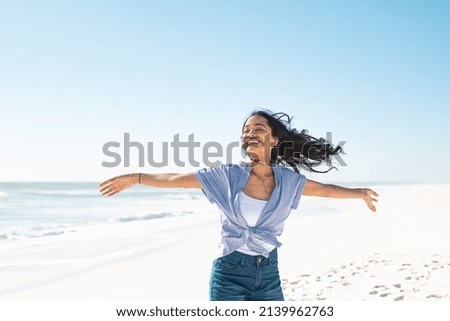 Healthy young woman standing on the beach with hands outstretched. Happy smiling latin woman with open arms feeling the breeze on seaside. Natural beauty woman with outstretched arms up dancing at sea