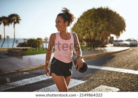 Smiling young African woman in a tank top and shorts listening to music on earphones while crossing the street carrying a yoga mat