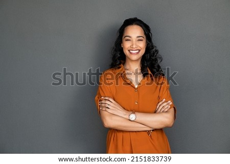Photo of Portrait of a young latin woman with pleasant smile and crossed arms isolated on grey wall with copy space. Beautiful girl with folded arms looking at camera against grey wall. Cheerful hispanic woman