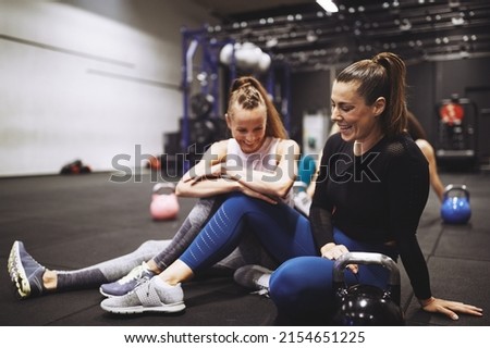 Two fit women in sportswear laughing while sitting together on the floor of a gym after an exercise class