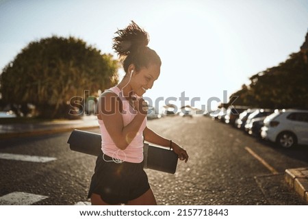 Smiling young African woman in sportswear listening to music on earphones while crossing a street carrying her yoga mat