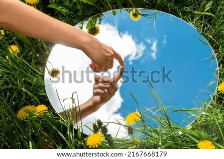 nature concept - hand touching sky reflection in round mirror on summer field