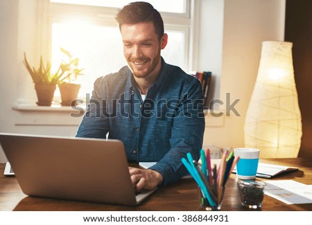 Successful entrepreneur smiling in satisfaction as he checks information on his laptop computer while working in a home office, sun flare behind
