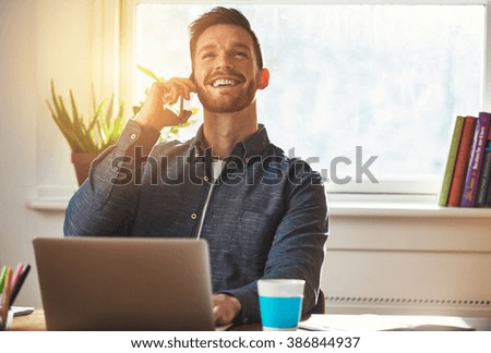 Confident entrepreneur chatting on a mobile phone while working at his desk looking up into the air with an elated smile of satisfaction