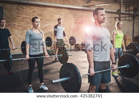 Serious muscular bearded man in shorts and gray tee shirt leading small group of young adults in barbell exercises for fitness training