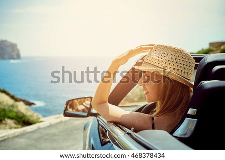 Belle jeune femme souriante et blonde avec chapeau et lunettes de soleil dans une voiture décapotable qui regarde de côté tandis qu