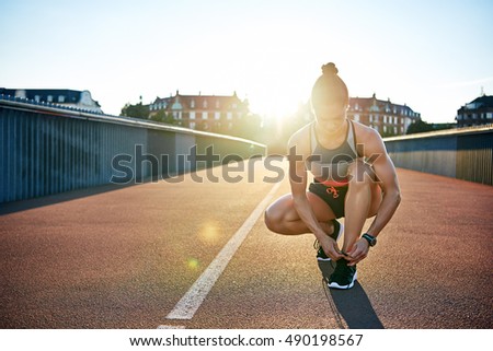 Muscular woman ties her running shoes on bridge as sun shines over her shoulder