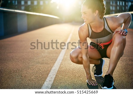 Woman kneels with one hand on pavement and smiles as she prepares to run down empty bridge