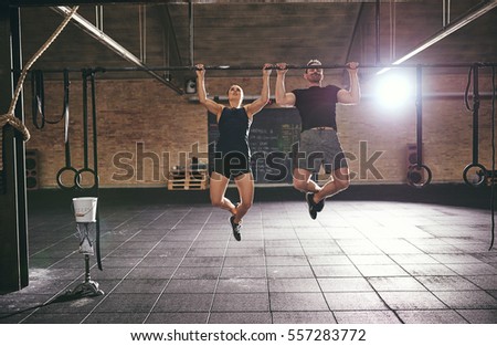 Front shot of two young athletic people dowing pull-ups together in spacious light gym. 