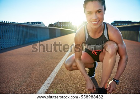 Muscular woman smiles at camera as she kneels on bridge to tie her shoes
