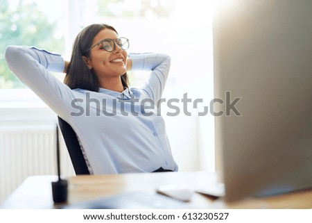 Woman with satisfied expression at desk. Arms up and folded behind her head in front of bright window.