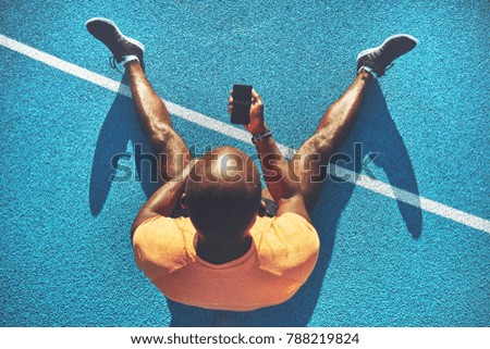 Young African male athlete in sportswear sitting alone on the lanes of a running track checking his running time on his smartphone