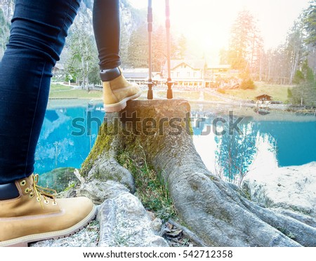 Young woman doing trekking with blue lake and chalet in background - Hiker enjoying summer season mountain sport - Healthy lifestyle and nature concept - Focus on left shoe walking poles - Warm filter