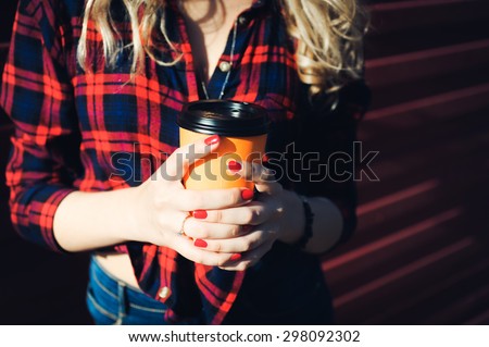 close-up portrait of a beautiful bright girl young sexy  blonde with red lips with curly hair with a cup of coffee  to go in a plaid shirt and denim shorts smiling and posing