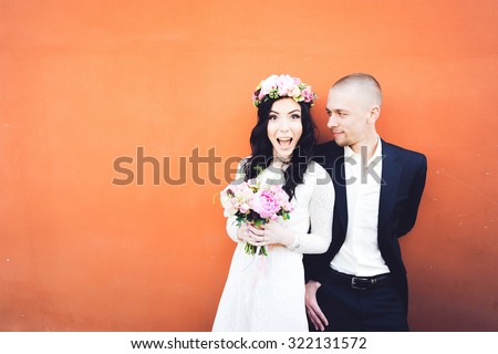 close-up portrait young beautiful stylish couple bride in a white dress with a bouquet of flowers in her hair and and groom in a suit on a background of an orange wall laughing and hugging each other