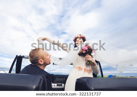 close-up portrait young beautiful stylish couple bride in a white dress with a bouquet of flowers in her hair and and groom in retro car on the way to their honeymoon