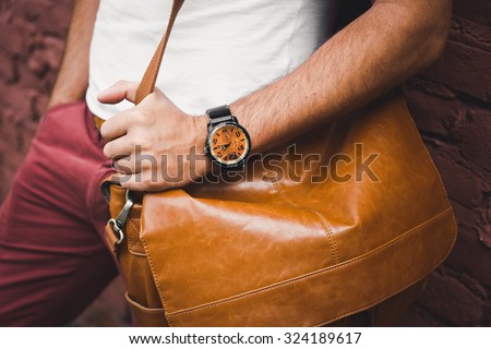 young guy with a beard and mustache  and white T-shirt  posing on the street  vintage man, fashion men, hipster street casual  leather bag and hours against the background of a brick wall