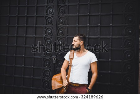 young guy with a beard and mustache  and white T-shirt  posing on the street  vintage man, fashion men, hipster street casual  leather bag and hours against the background of a brick wall