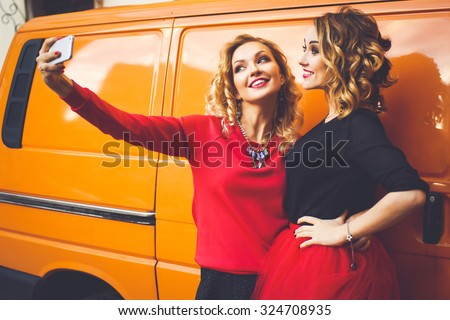 Portrait of young beautiful stylish girl friends on the streets in a red blouse and red skirt on a sunny day on a background of orange cars posing and smiling for Life Style do self photo on the phone