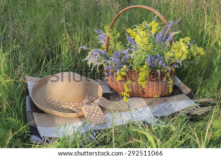 Still life with a blanket, a basket of flowers and women\'s hat. Summer in the meadow.