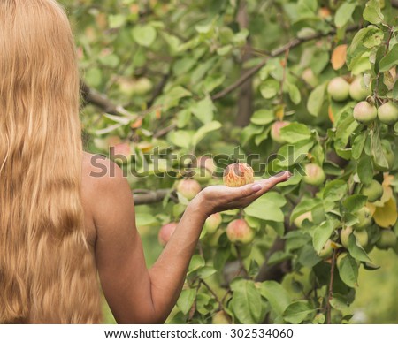 A girl with long red hair standing with his back to Apple orchard and holding an Apple. Eve with the Apple in his hand.