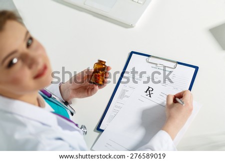 Beautiful young doctor sitting in front of working table , holding container with pills and writing prescription. Medical concept. Handsome female doctor distracted from paperwork looking up