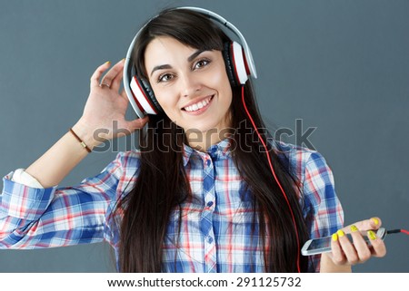 Beautiful dark haired smiling woman wearing headphones, holding phone and listening music on grey background