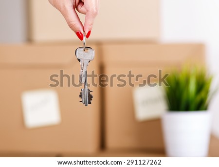 Female hand holding keys over pile of brown cardboard boxes with house or office goods background. Moving to new place of living concept.
