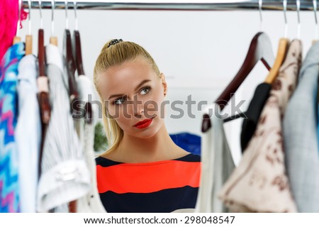 Thoughtful beautiful blonde woman standing near wardrobe rack full of clothes and choosing dress. Shopping and consumerism or stylist concept. Nothing to wear and hard to decide concept