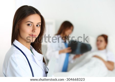 Beautiful female medicine doctor looking in camera in front of patient lying in bed and communicating with traumatologist. Radiology or traumatology medical concept. Medical care or insurance concept