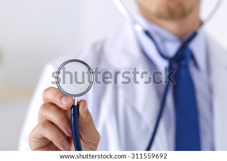 Male medicine doctor hand holding stethoscope head closeup in front of his chest. Medical help or insurance concept. Physician is ready to examine patient and help. Treatment and patient care concept