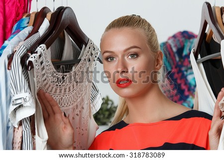 Beautiful thoughtful blonde woman standing inside wardrobe rack full of clothes suffering with choice. Shopping and consumerism or stylist concept. Nothing to wear and hard to decide concept