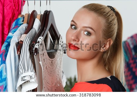 Beautiful smiling blonde woman standing inside wardrobe rack full of clothes suffering with choice. Shopping and consumerism or stylist concept. Nothing to wear and hard to decide concept