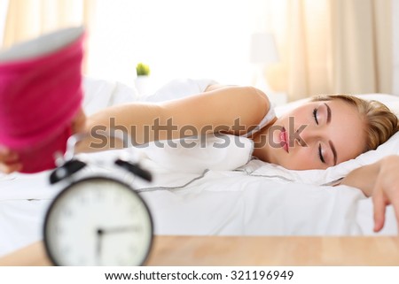 Sleepy young beautiful woman trying kill alarm clock with pink slipper. Early wake up, not getting enough sleep, getting work concept. Female stretching hand to ringing alarm willing turn it off