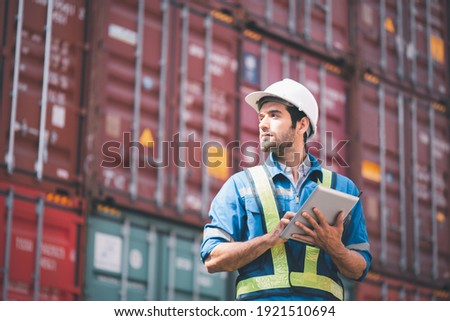 Engineer or foreman holding tablet and wears PPE looking at left side to checking inventory or job details with cargo container background. Engineering site and working with technology concept.