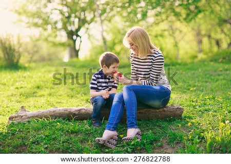 happy mother and her young son sitting on a tree in the park and eat a big red apple