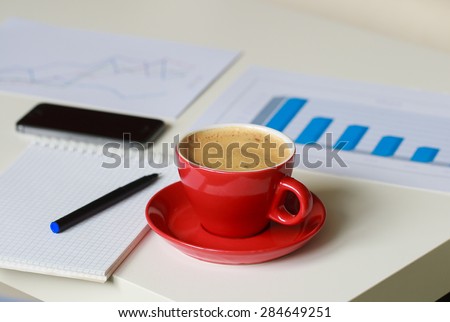 a red cup of coffee, business diagrams, mobile phone, note book and black pen on the table
