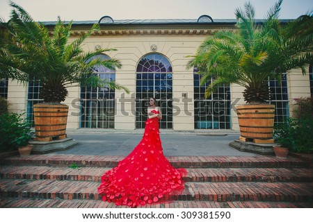 beautiful girl in long red evening gown dress is standing near the palm trees in city park