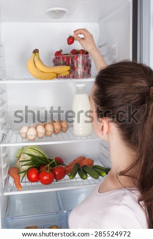 Young cheerful woman taking a strawberry  from refrigerator,  healthy eating concept